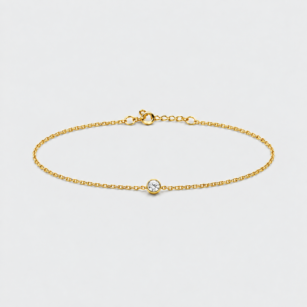 A delicate gold chain bracelet with a small bezel-set clear gemstone, displayed on a light gray background.
