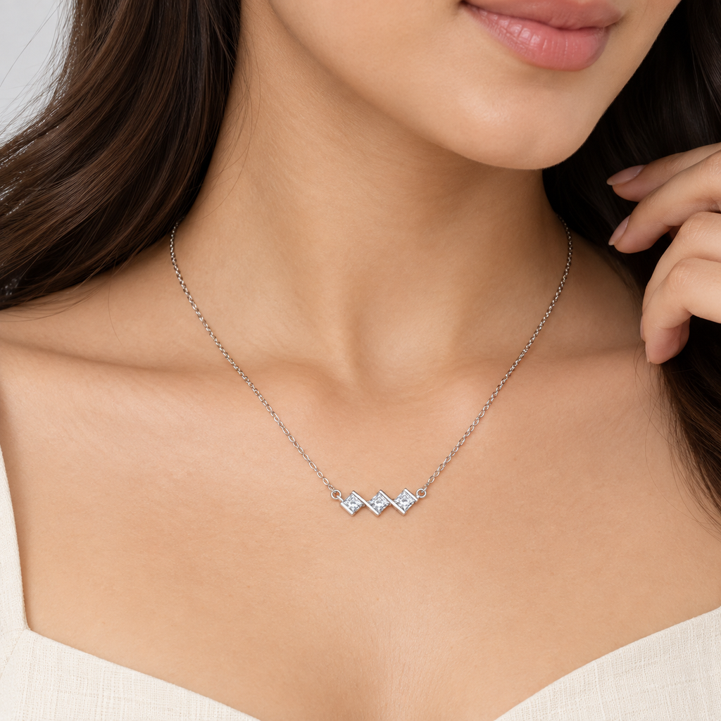 A close-up of a woman wearing a delicate silver chain necklace with three small, clear, diamond-cut geometric charms arranged in a zigzag pattern.