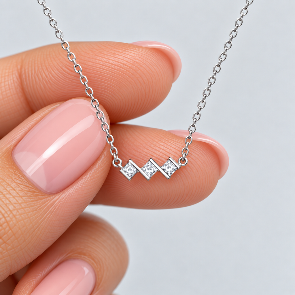 A close-up of a hand with pink manicured nails holding a delicate silver chain necklace with three small diamond-shaped clear gemstone clusters on a light gray background.