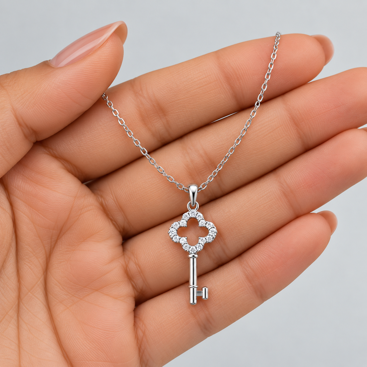 A silver key-shaped pendant necklace with a four-petal flower design set with small clear stones, held in the palm of a hand against a light gray background.