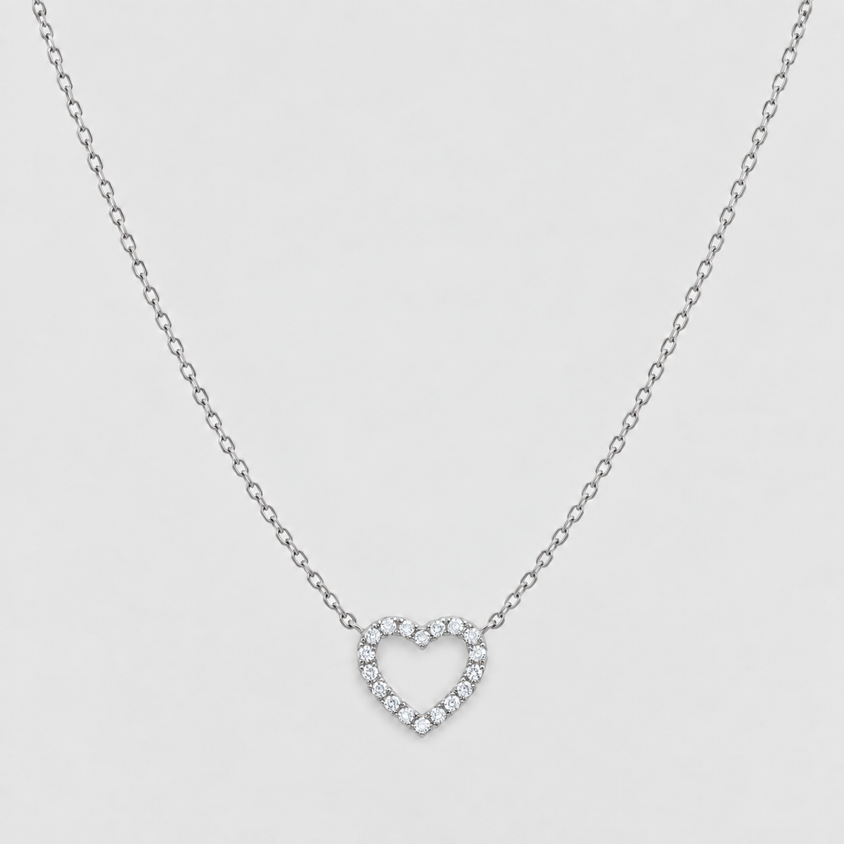 A delicate silver chain necklace featuring a small open heart-shaped pendant encrusted with clear stones, presented on a light gray background.