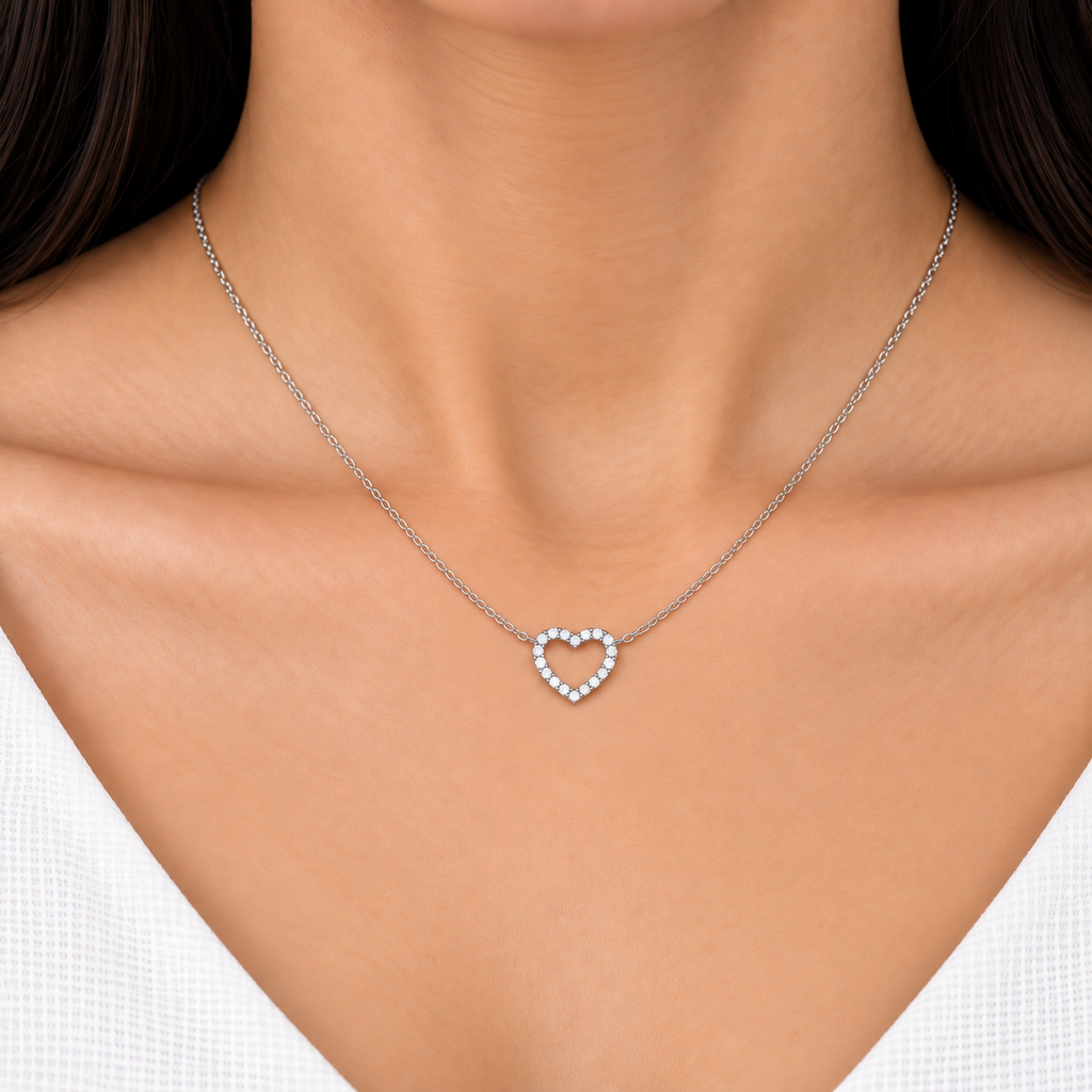 A close-up of a model's neck and chest wearing a delicate silver chain necklace with a small heart-shaped pendant set with clear stones.