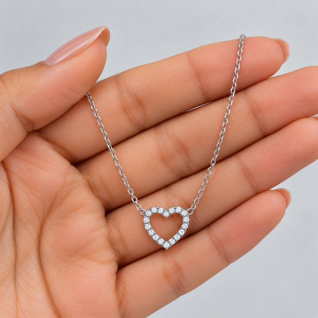 A delicate silver heart pendant necklace adorned with small clear stones, held in the palm of a hand against a soft light gray background.