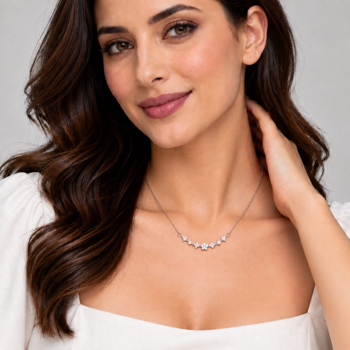 A woman with long wavy brown hair models a delicate silver necklace featuring a curved row of small star-shaped clear gemstones. She is wearing a white off-the-shoulder top against a neutral light gray background.