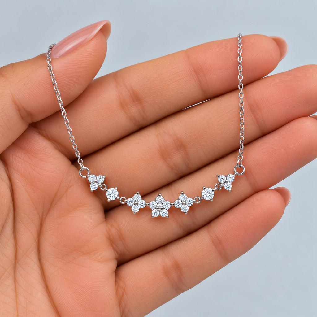 A delicate silver chain necklace with five small flower-shaped clusters of clear stones, held across an open hand against a light gray background.