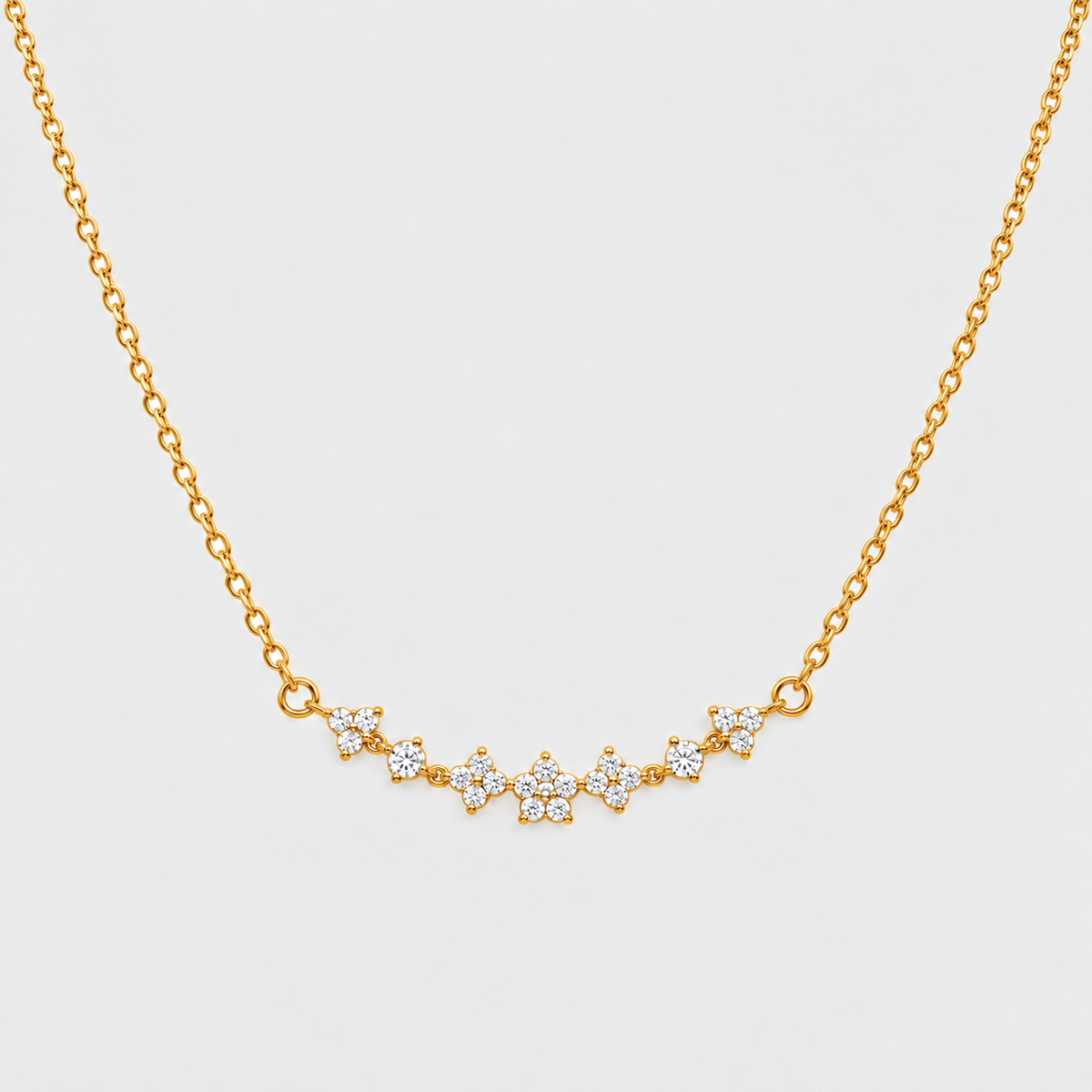 A delicate gold chain necklace featuring a curved bar of small floral clusters set with clear stones, displayed against a light gray background.