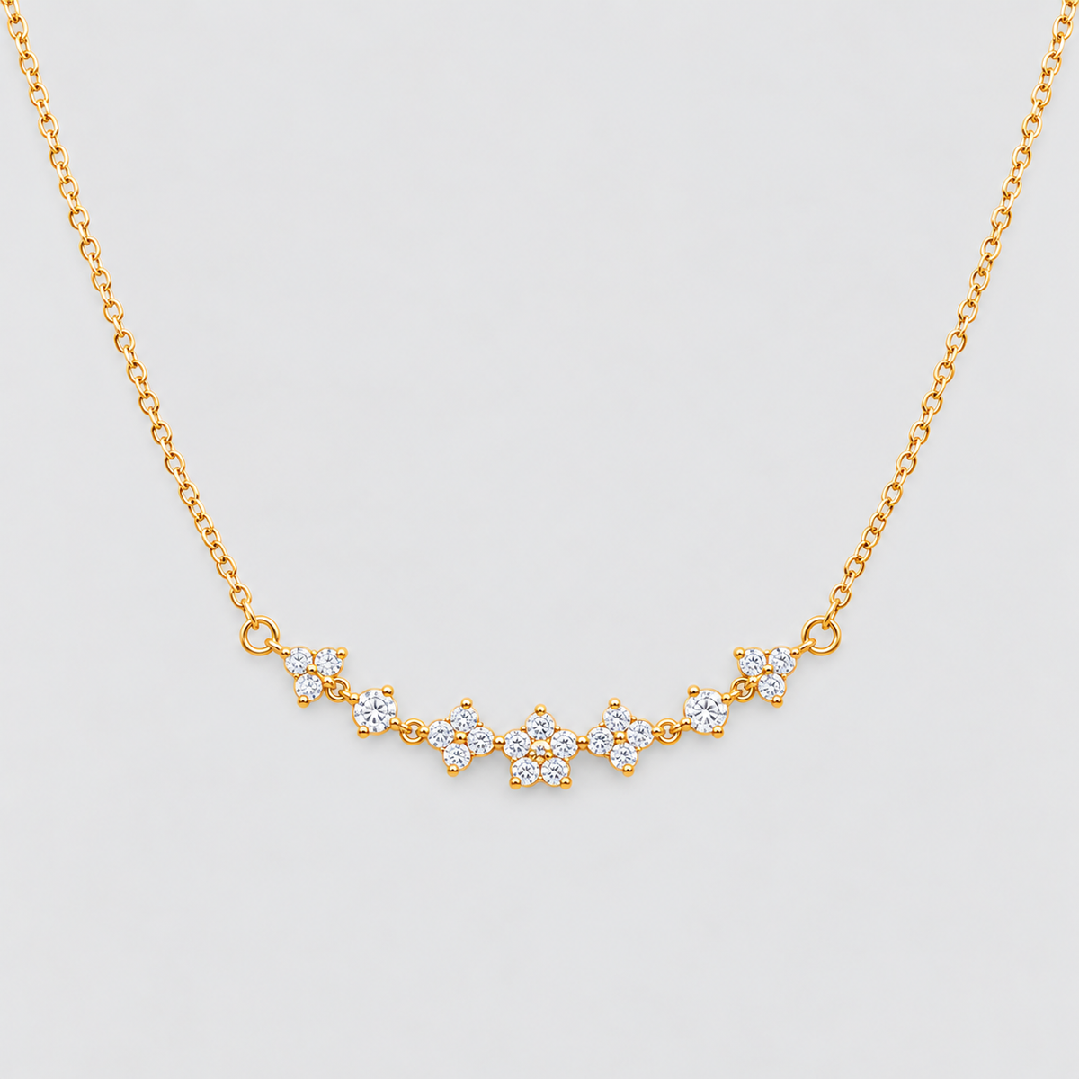 A delicate gold chain necklace featuring a curved bar of small flower-shaped clusters set with clear stones, displayed against a light gray background.