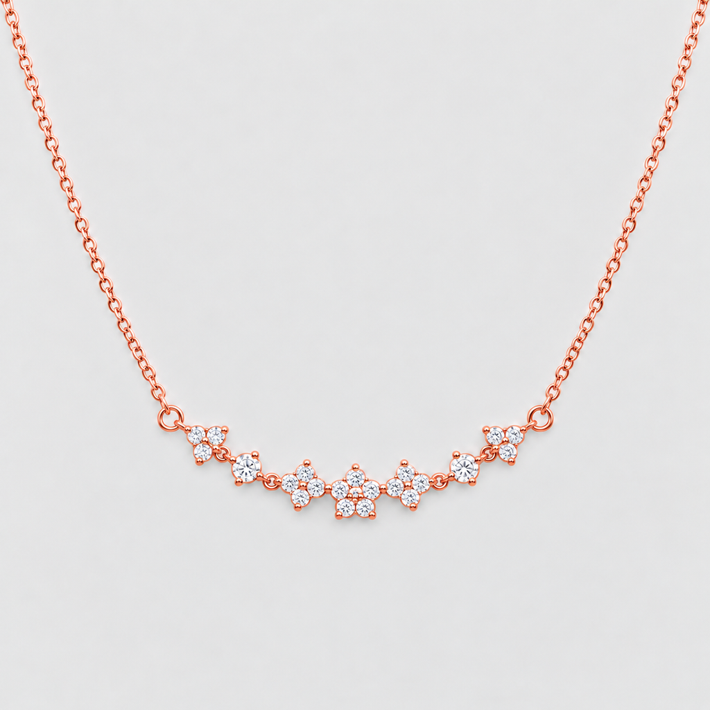 A delicate rose gold chain necklace featuring a curved bar of small floral clusters set with clear stones, displayed against a light gray background.