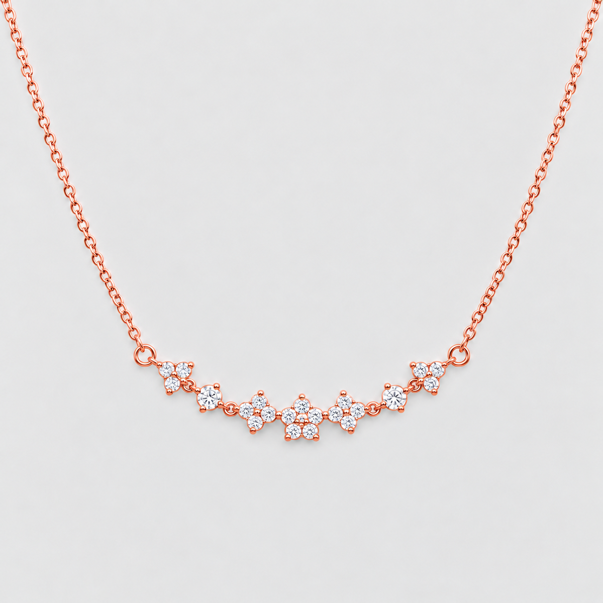 A delicate rose gold chain necklace featuring a curved bar of small floral clusters set with clear stones, displayed against a light gray background.
