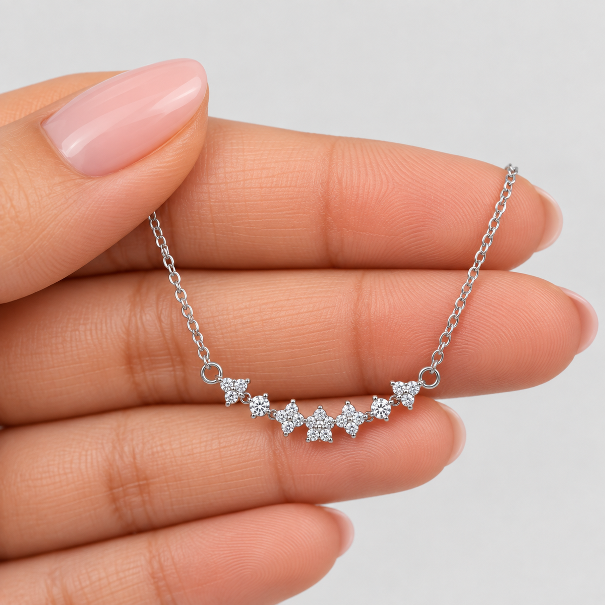 A delicate silver chain necklace with a curved row of small flower-shaped clusters of clear stones, held between fingers against a light gray background.