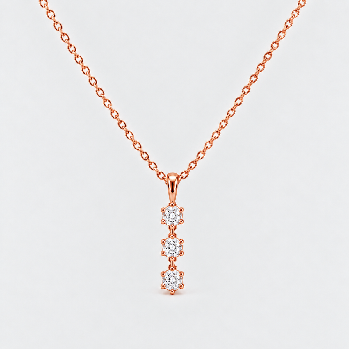 A rose gold necklace with a delicate chain and a vertical pendant featuring three small round-cut clear gemstones in prong settings, shown on a light gray background.