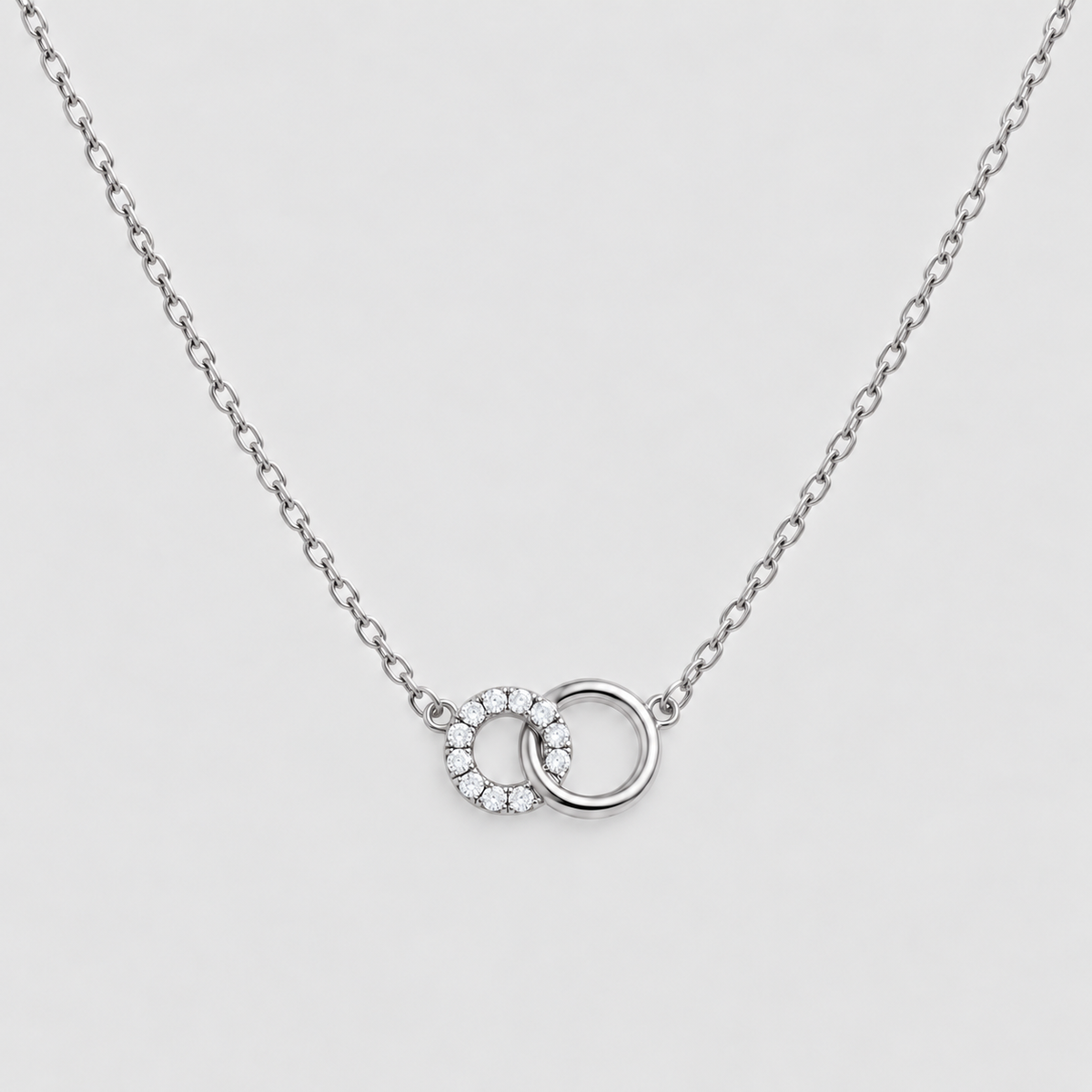 A silver chain necklace featuring a double interlocking ring pendant, with one ring detailed with small clear stones, set against a light gray background.