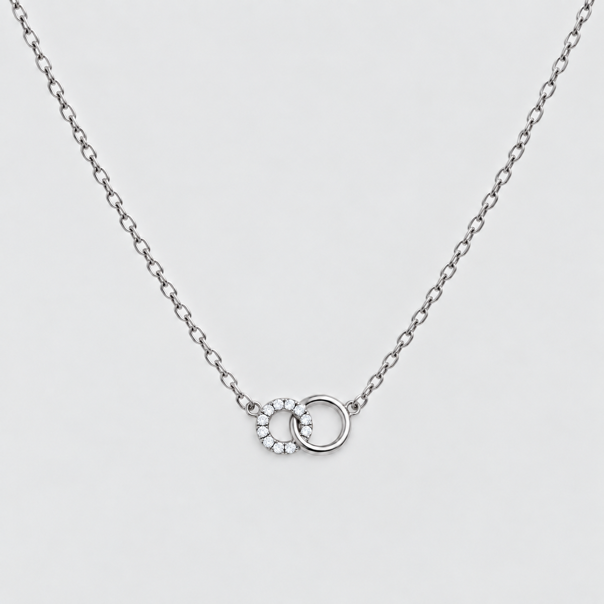 A delicate silver chain necklace featuring a pendant with two interlocking rings, one adorned with small clear stones, set against a light gray background.