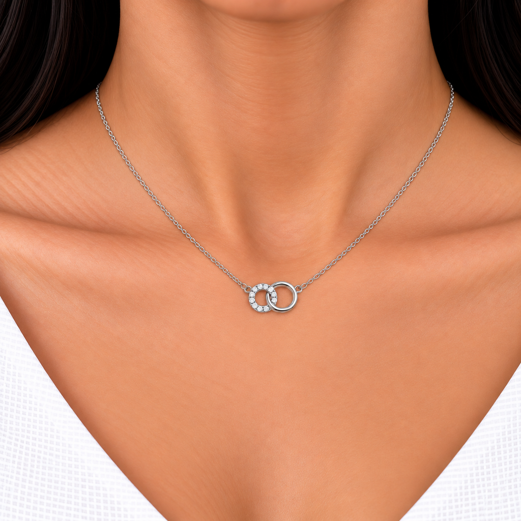 A close-up of a model's neck and chest wearing a delicate silver chain necklace with a double interlocking ring pendant, one of which is set with small clear stones.