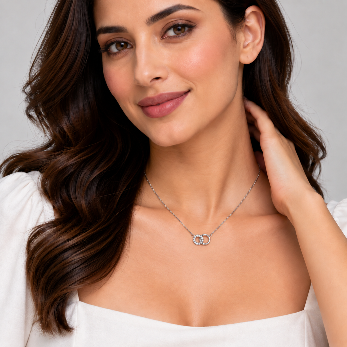 A woman with long wavy brown hair models a delicate silver necklace featuring a curved row of small star-shaped clear gemstones. She is wearing a white off-the-shoulder top against a neutral light gray background.