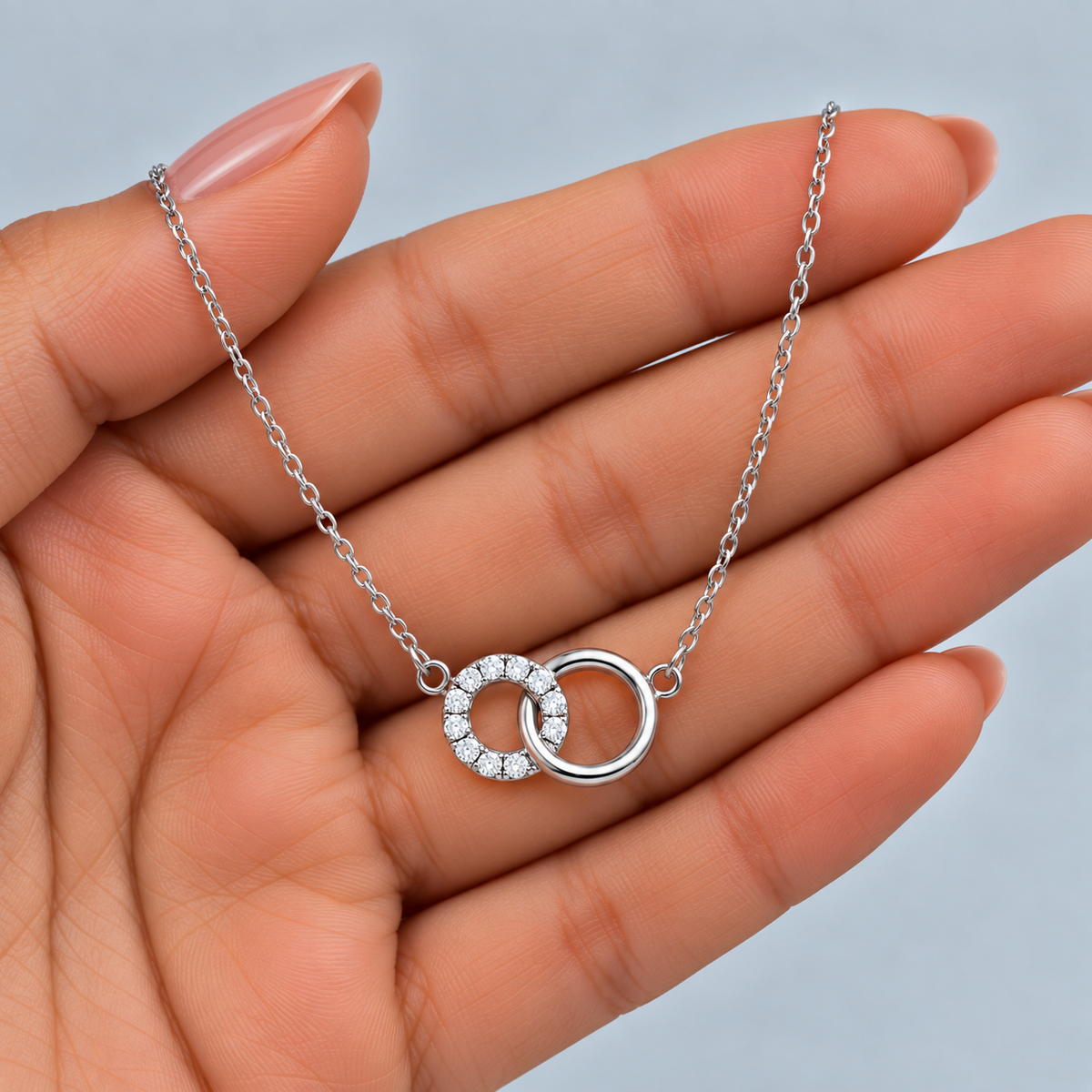 A silver necklace with two interlocking ring pendants, one of which is set with small clear stones, held in a person's hand against a light gray background.