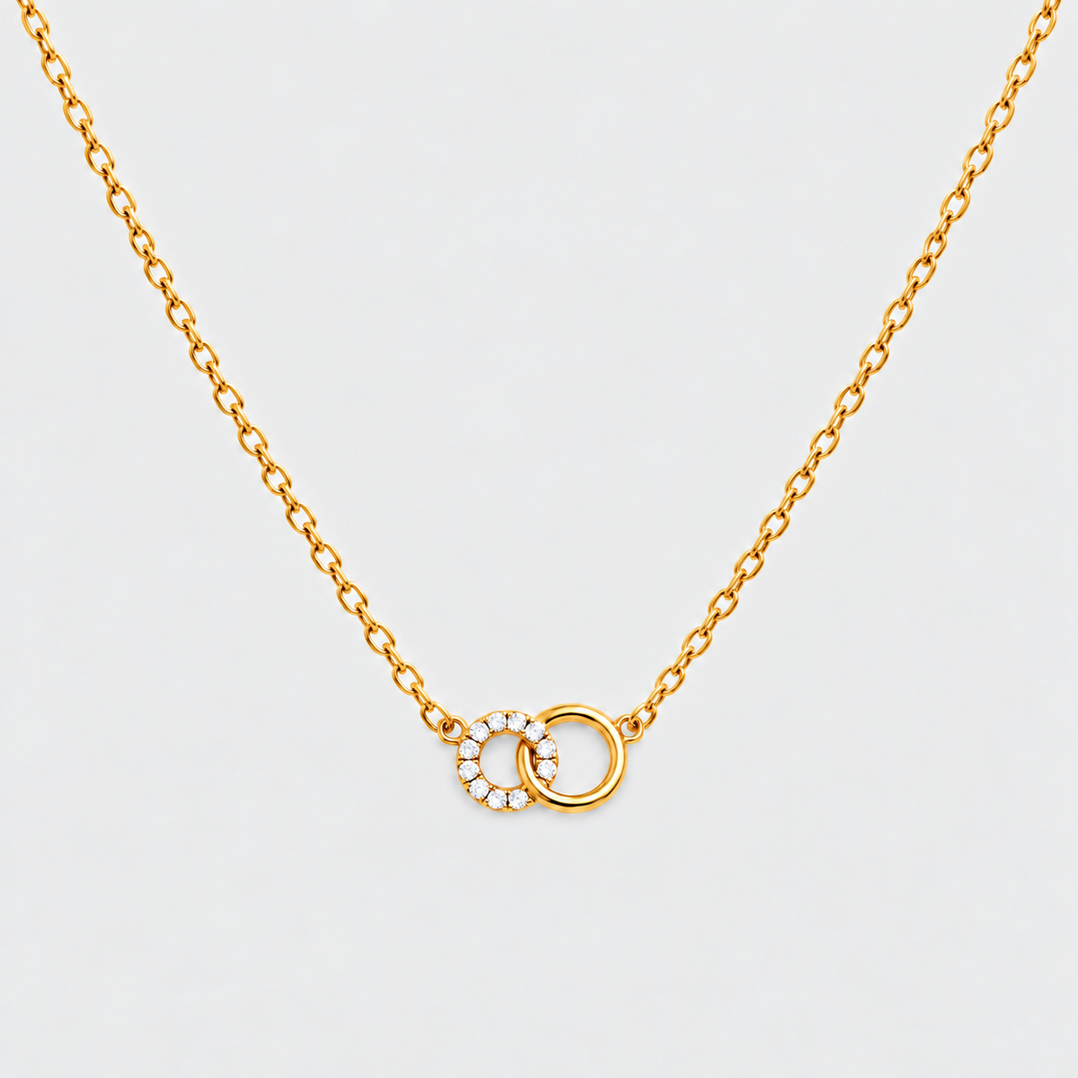 A delicate gold chain necklace featuring two interlocking ring pendants, one adorned with small clear stones, set against a light gray background.