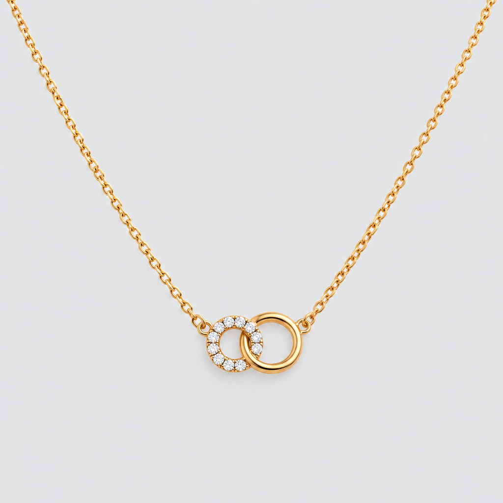 A delicate gold chain necklace featuring two interlocking circular pendant elements, one of which is adorned with small clear stones, set against a clean light gray background.