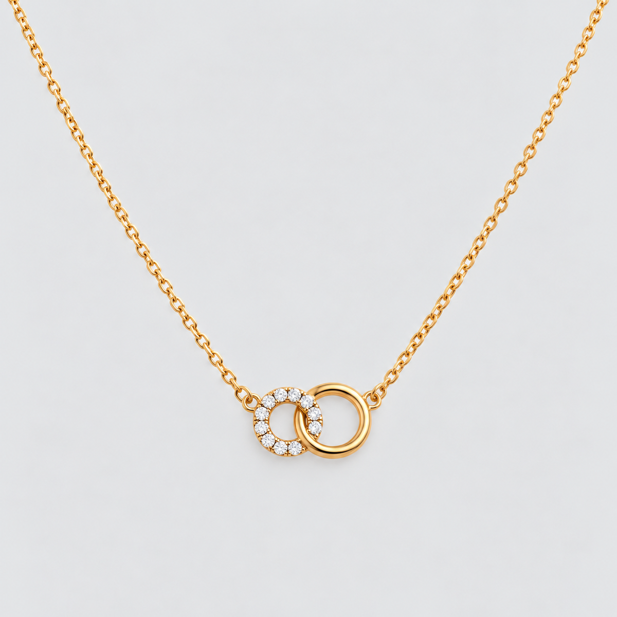 A delicate gold chain necklace featuring two interlocking circular pendant elements, one of which is adorned with small clear stones, set against a clean light gray background.