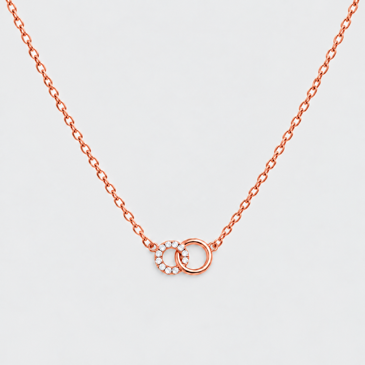 A delicate rose gold chain necklace featuring two interlocking ring pendants, one of which is adorned with small clear stones, set against a light gray background.