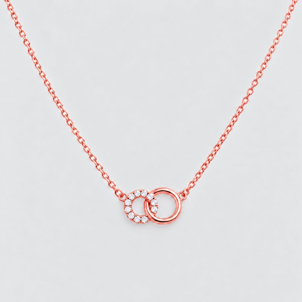 A delicate rose gold chain necklace featuring two interlocking rings at the center, one adorned with small clear stones, set against a light gray background.