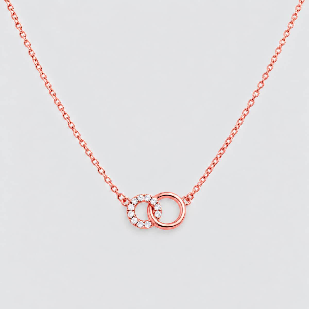 A delicate rose gold chain necklace featuring two interlocking rings at the center, one adorned with small clear stones, set against a light gray background.