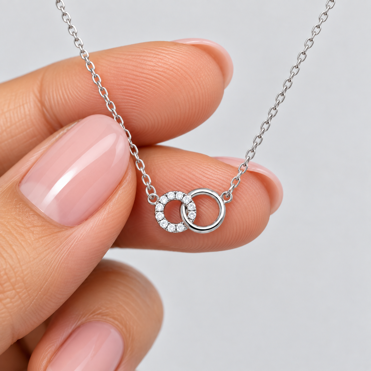 A close-up of a hand with pink manicured nails holding a delicate silver chain necklace with a double interlocking ring pendant, one of which is set with small clear stones, against a light gray background.