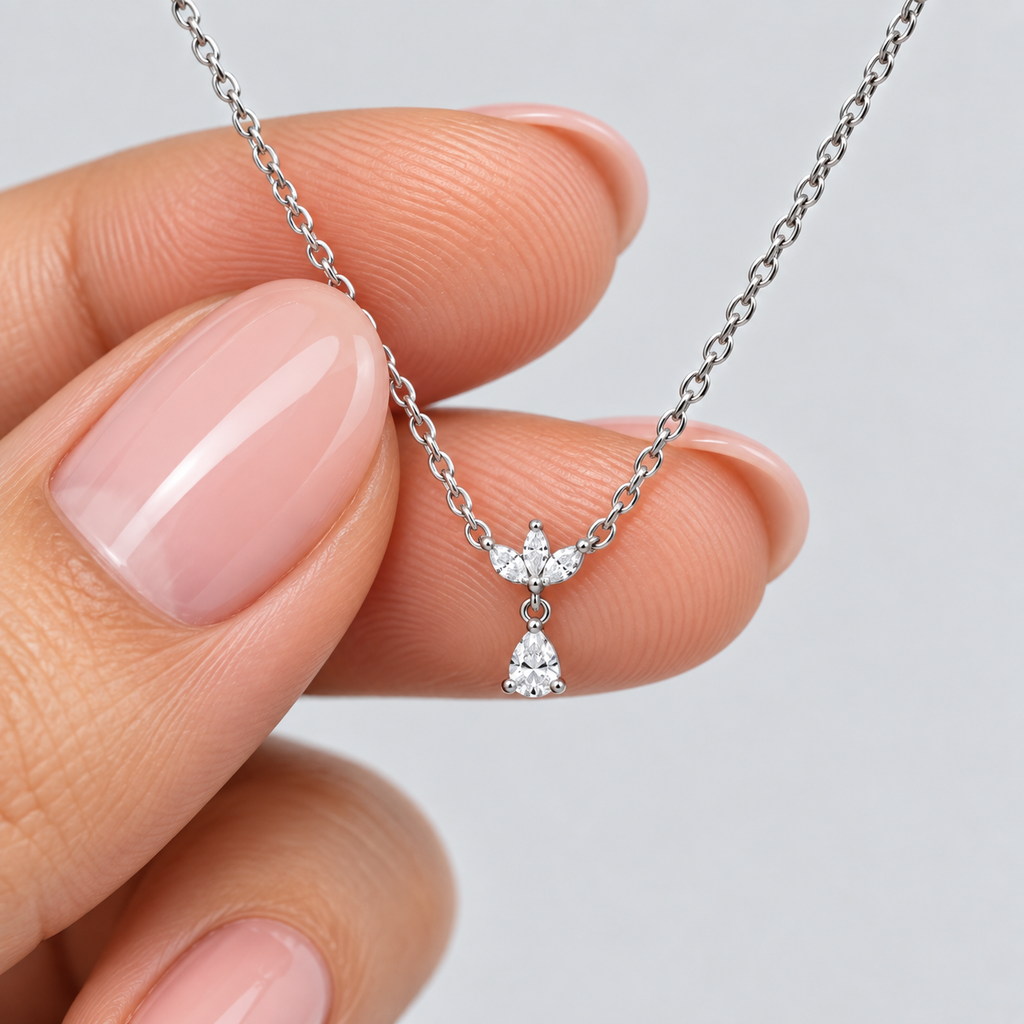 A close-up of a delicate silver chain necklace with a small winged pendant and a dangling teardrop-shaped clear gemstone, held between two fingers against a light gray background.