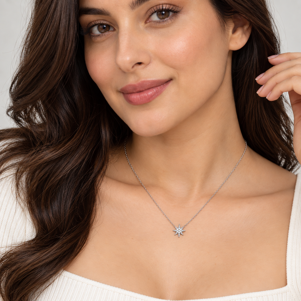 A close-up of a woman wearing a delicate silver chain necklace with a small starburst pendant, styled with a white ribbed top against a soft neutral background.