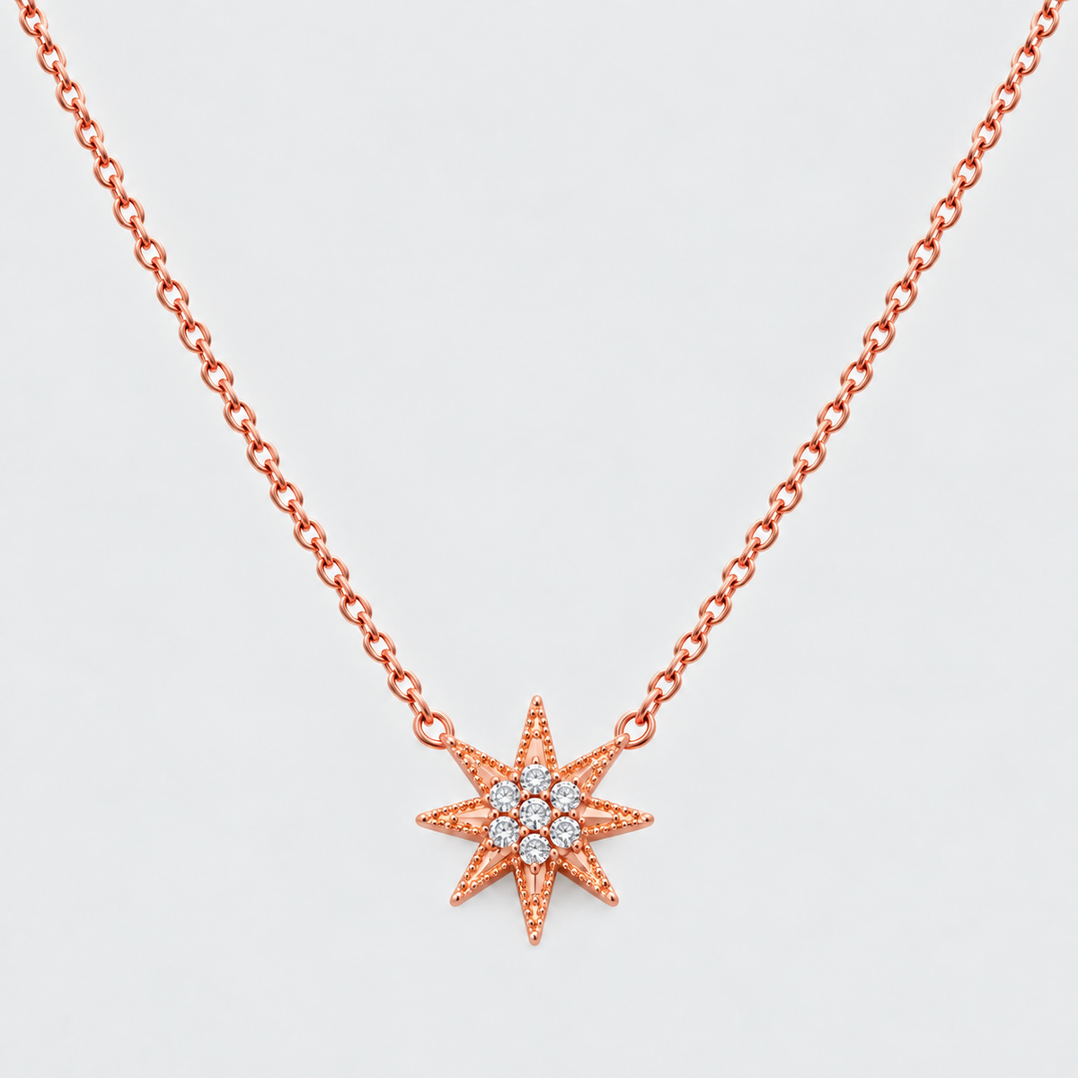 A rose gold necklace with a starburst pendant featuring a central cluster of small clear gemstones, shown against a light gray background.