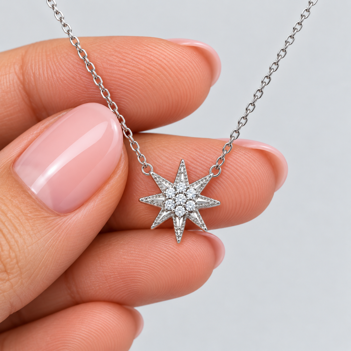 A silver-toned eight-pointed star pendant necklace with small clear stones, held between a person's fingers against a light gray background.