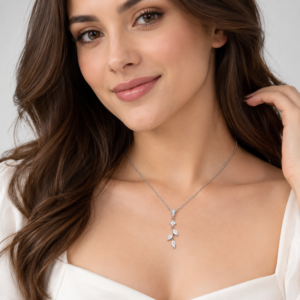 A woman with long brown hair wearing a delicate silver-toned leaf-shaped pendant necklace with clear stones, paired with a white top against a light gray background.