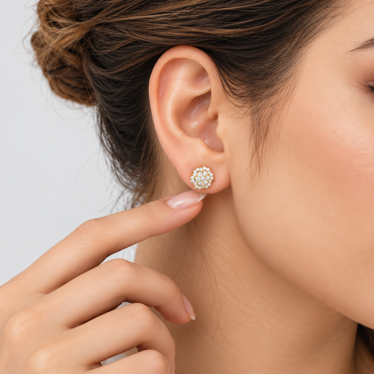 A close-up of a woman's ear wearing a small round stud earring with a halo of clear stones in a yellow metal setting, with her finger touching the lobe. The background is a clean, light gray studio backdrop.