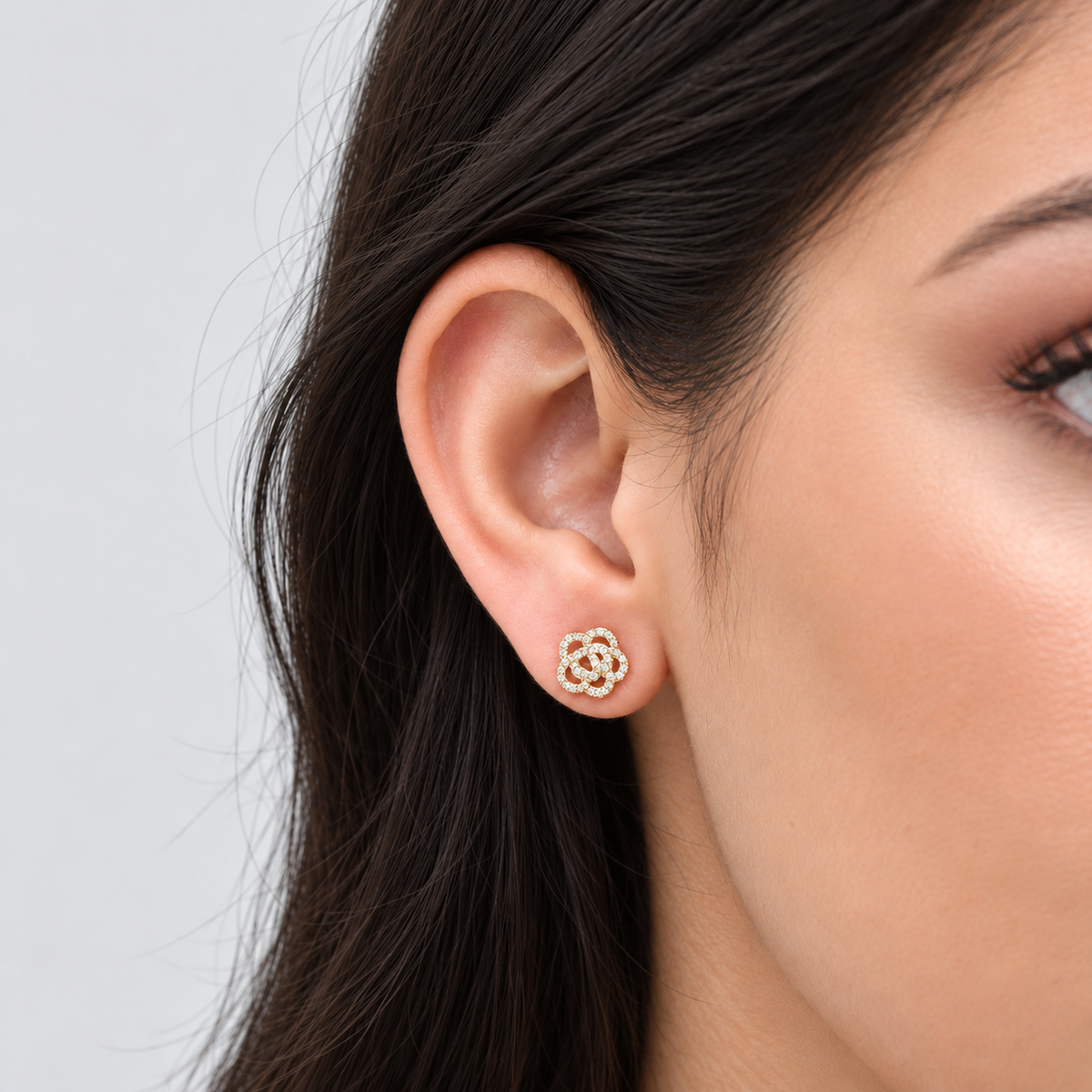A close-up of a woman's ear wearing a small gold stud earring with a floral or lotus-like openwork design set with tiny clear stones.