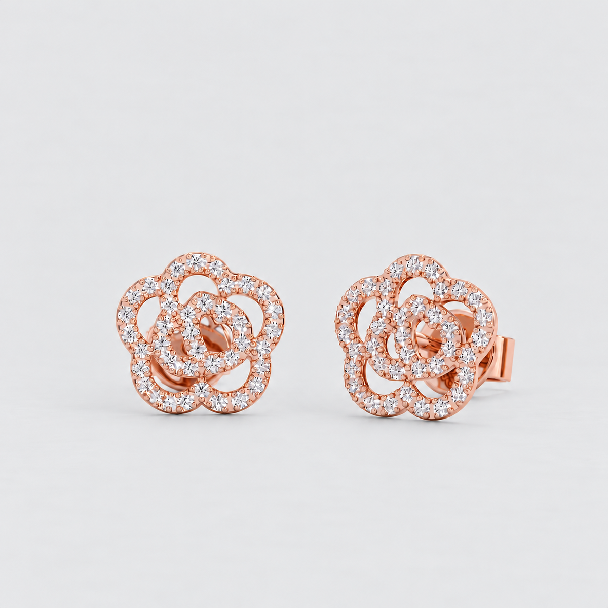 A pair of rose gold stud earrings with a multi-layered flower design paved with small clear stones, set against a clean light gray background.