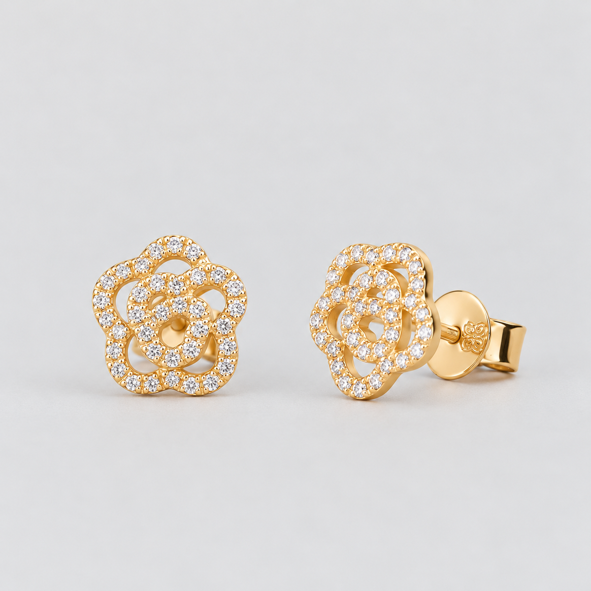 A pair of gold flower-shaped stud earrings paved with small clear stones, shown on a light gray background with one earring facing forward and the other angled to show the post and backing.