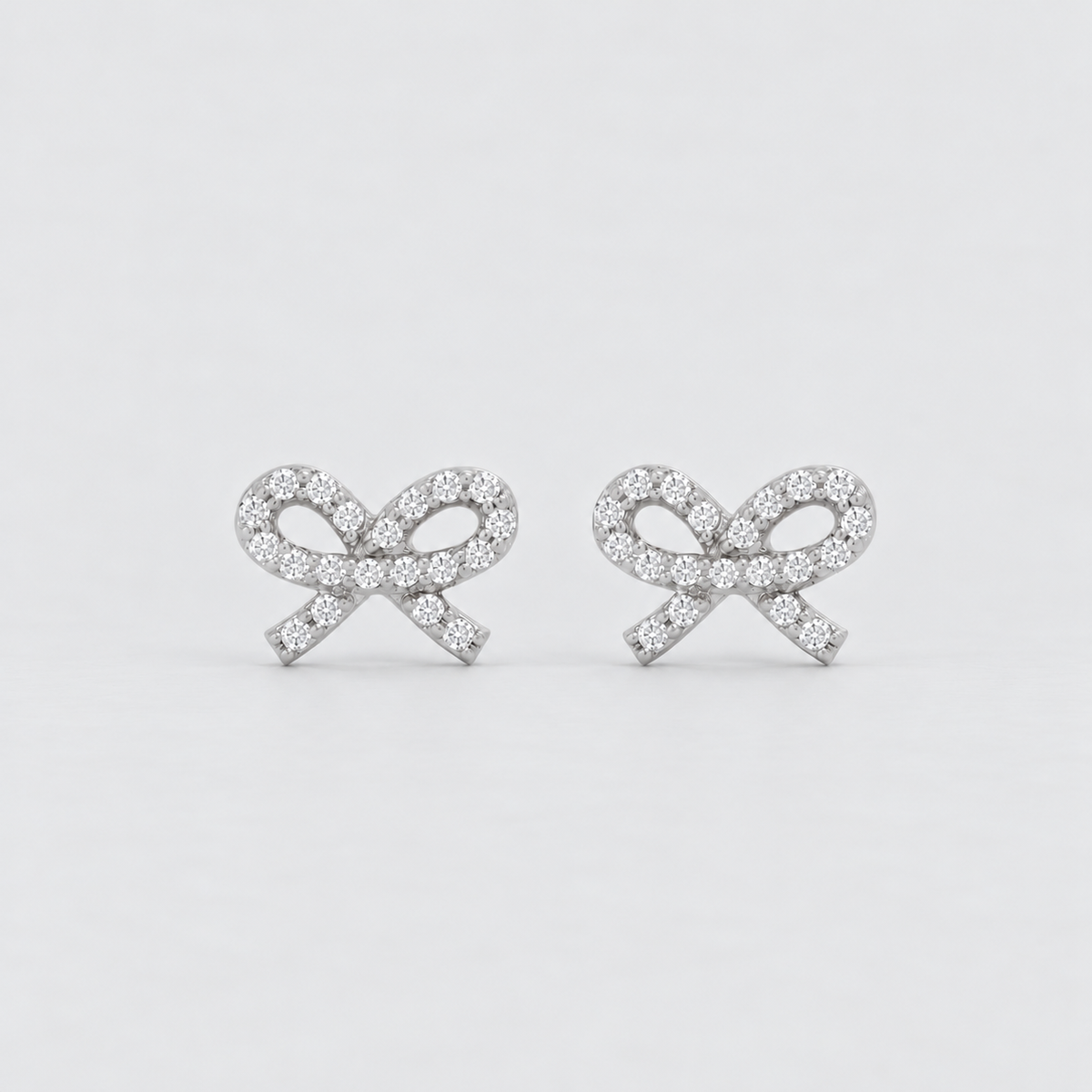 A pair of silver bow stud earrings paved with small clear gemstones, set against a clean light gray background.