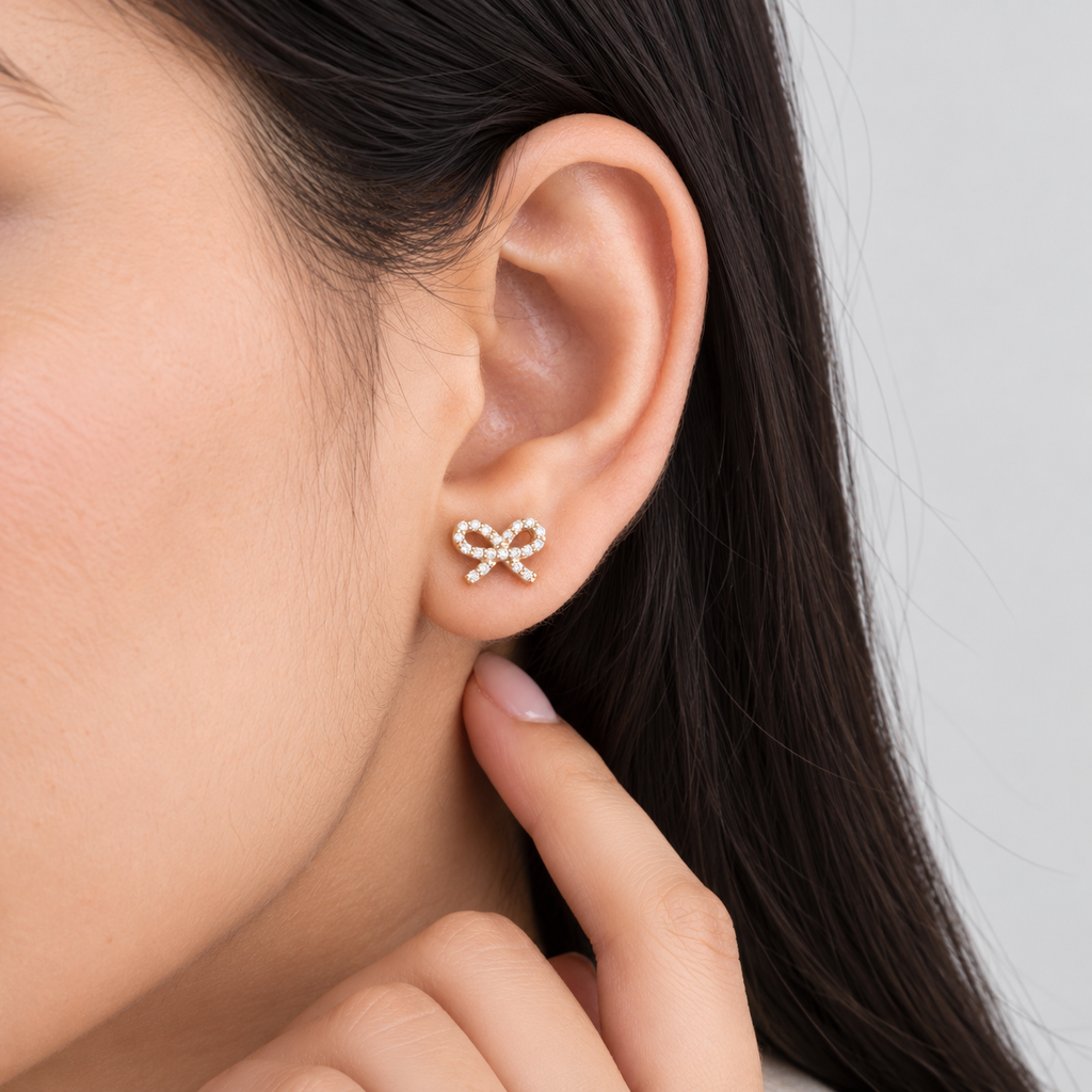 A close-up of a woman's ear wearing a small bow-shaped stud earring set with clear stones, with her finger touching her neck against a light gray background.