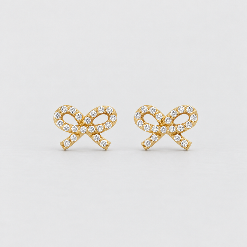 A pair of gold bow-shaped stud earrings paved with small clear gemstones, set against a clean light gray background.