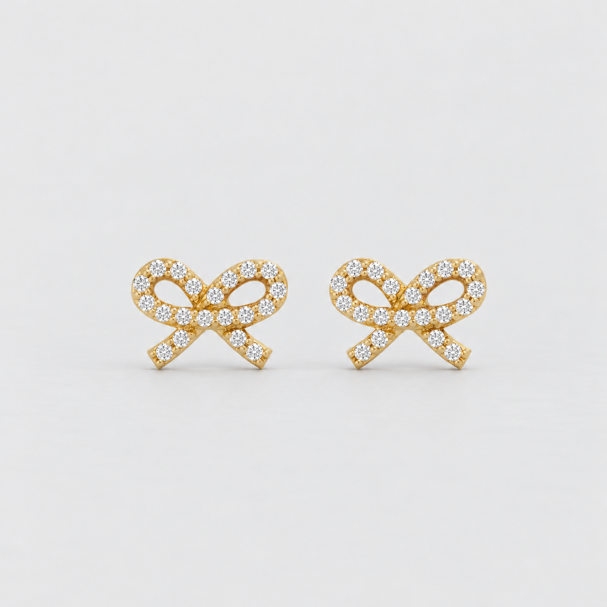 A pair of gold bow-shaped stud earrings paved with small clear gemstones, set against a clean light gray background.
