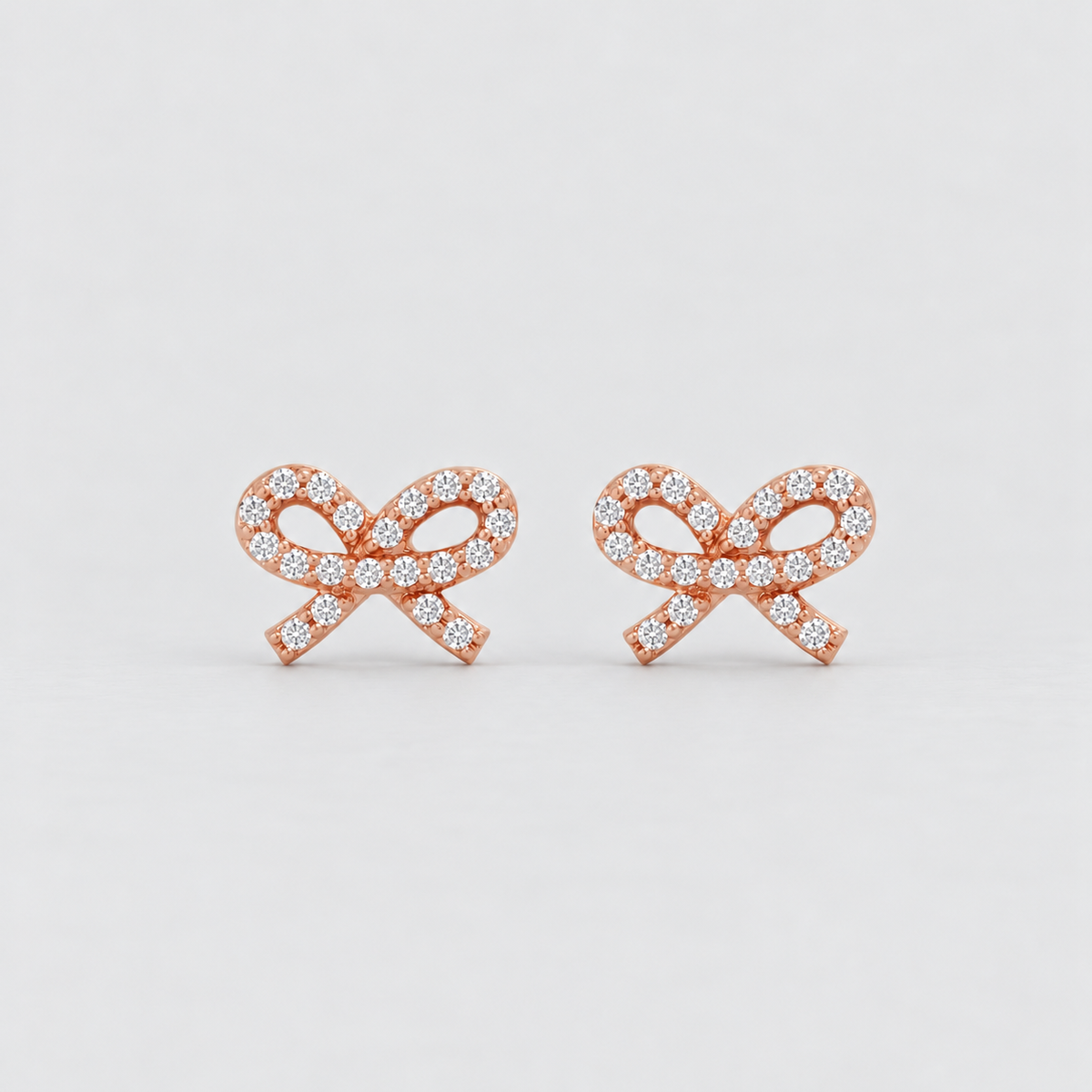 A pair of rose gold bow-shaped stud earrings paved with small clear gemstones, set against a clean light gray background.