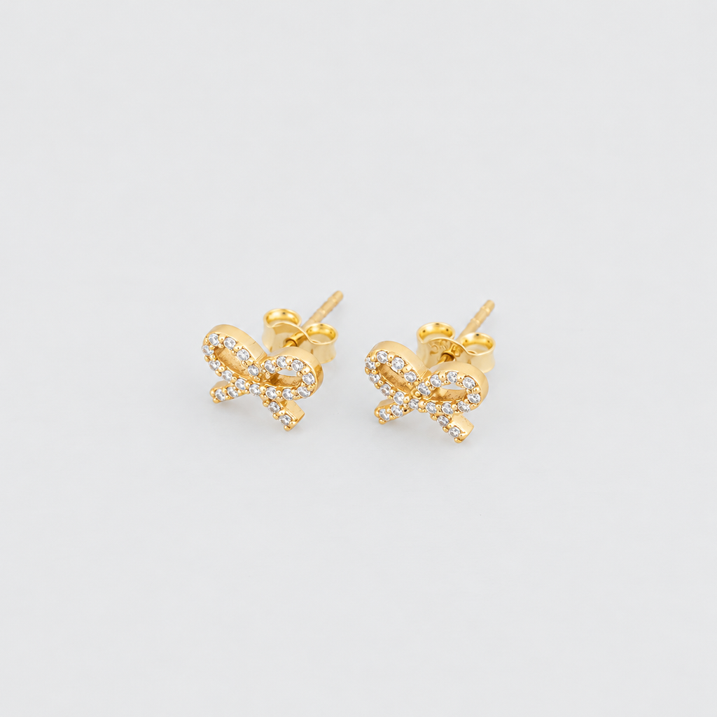 A pair of gold bow stud earrings adorned with small clear pavé stones, shown on a light gray background.