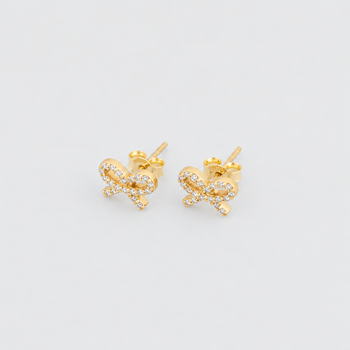 A pair of gold bow stud earrings adorned with small clear pavé stones, shown on a light gray background.