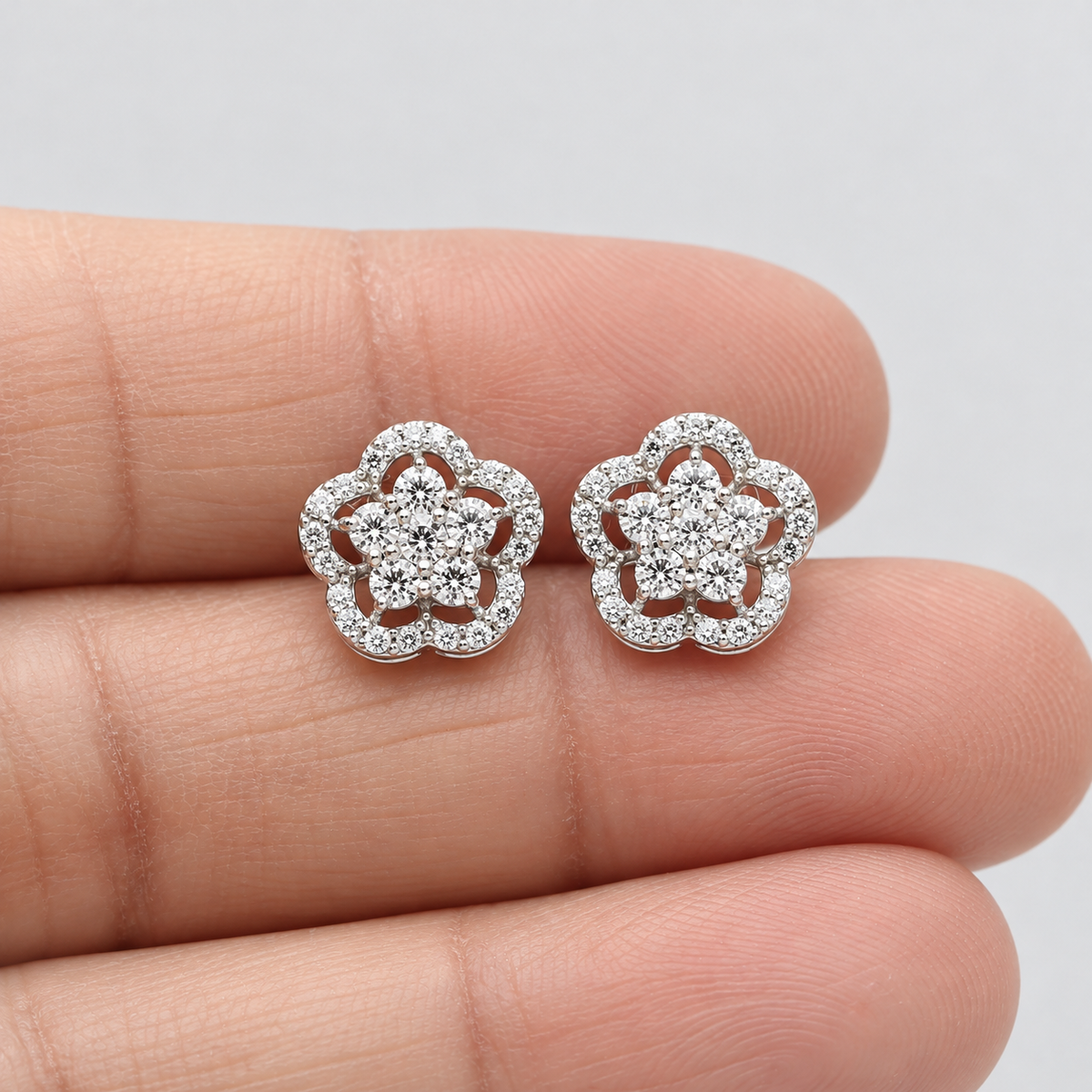 Pair of flower-shaped diamond cluster stud earrings in white gold held on a person's hand.