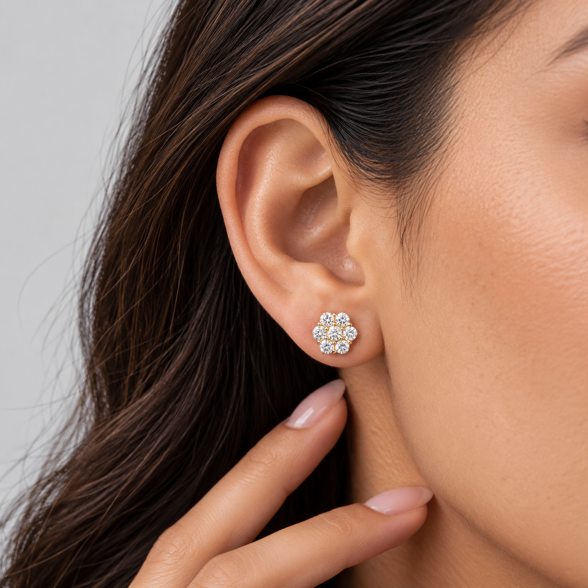 A close-up shot of a woman wearing a cluster diamond stud earring on her earlobe. The earring features multiple small round diamonds arranged in a flower-shaped cluster pattern.
