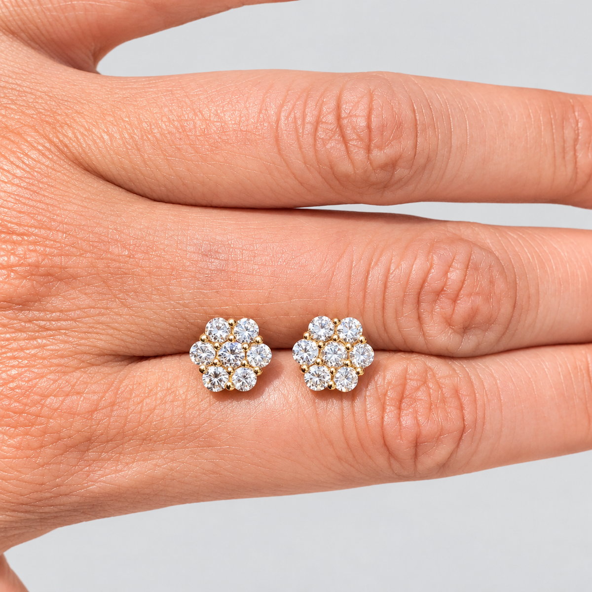 Two cluster diamond stud earrings shaped like small flowers, held against the back of a person's hand to show scale.