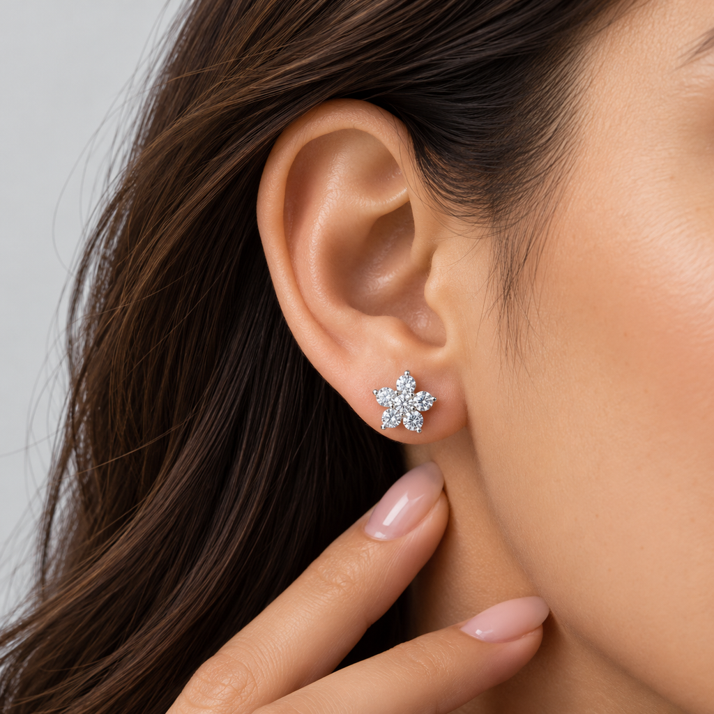 A close-up of a model's ear wearing a small silver flower-shaped stud earring with clear stones, with her hand resting near her neck.