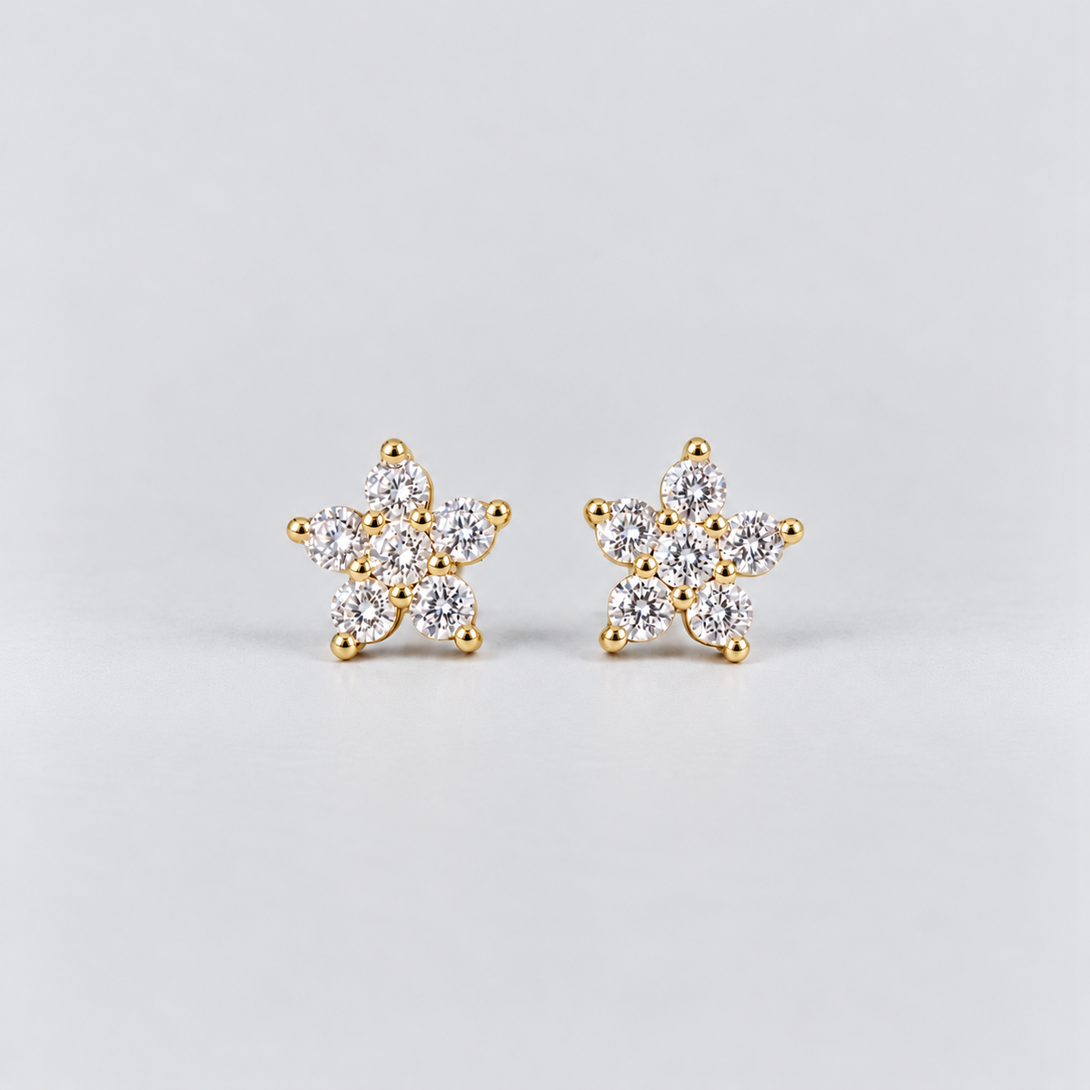 A pair of gold flower-shaped stud earrings set with clear round-cut gemstones, photographed on a clean light gray background.