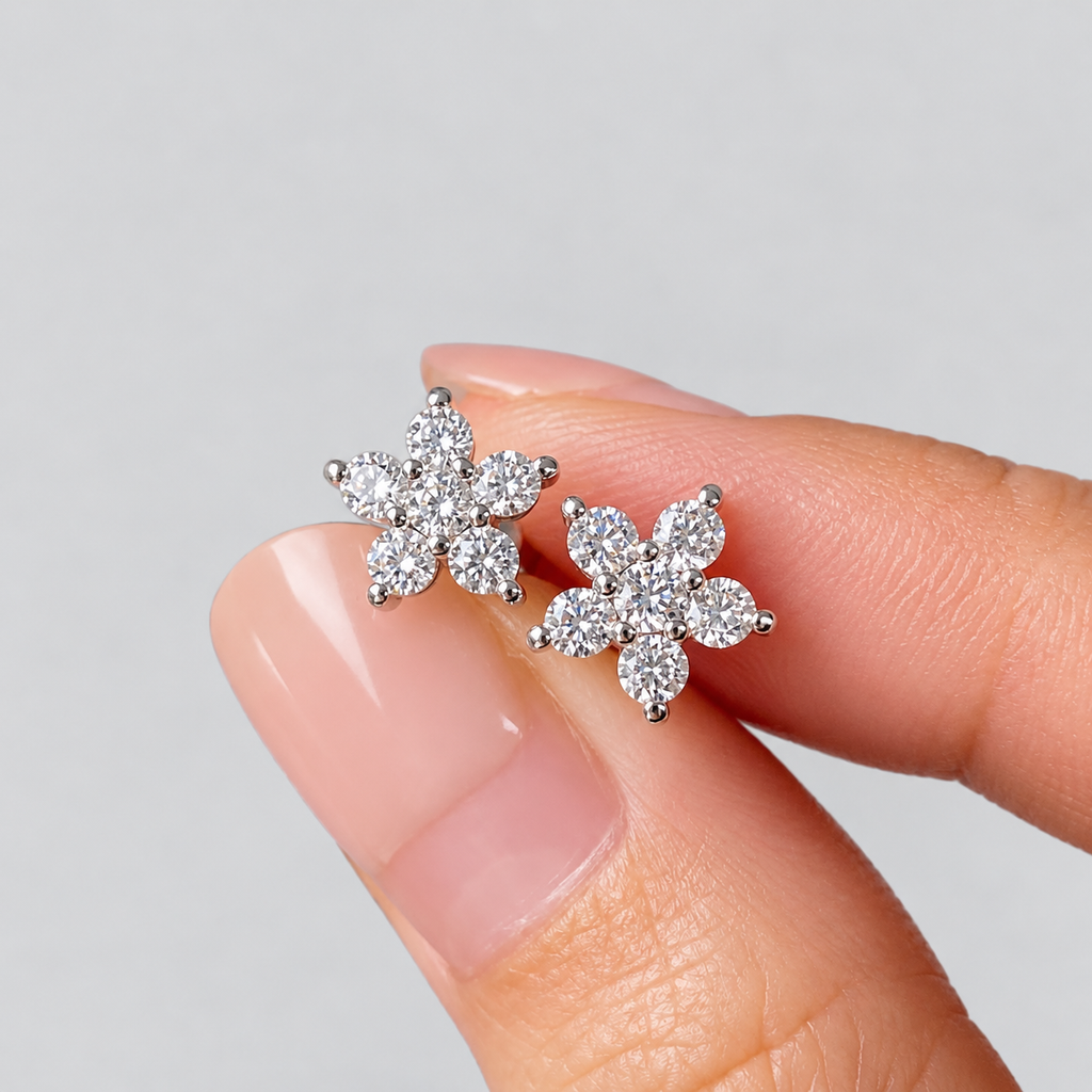 A pair of flower-shaped stud earrings with clear gemstones in a silver setting, held between two fingers against a light gray background.