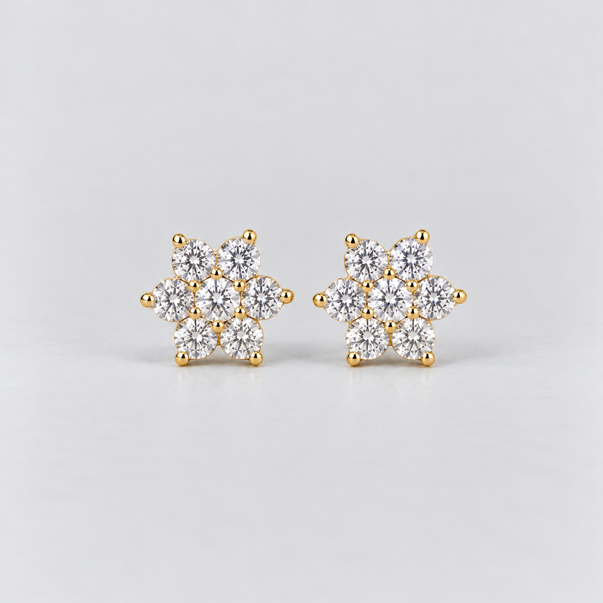 A pair of gold stud earrings with a snowflake-like cluster of clear round-cut gemstones, set against a clean light gray background.